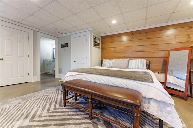 bedroom with concrete flooring, wood walls, a drop ceiling, and ensuite bathroom