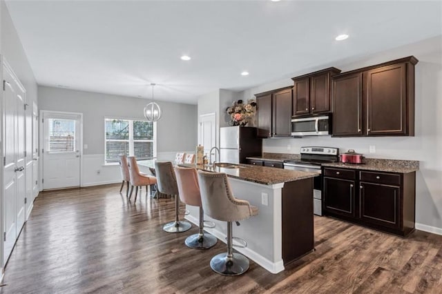 kitchen with dark wood-style flooring, a kitchen island with sink, dark stone counters, dark brown cabinets, and appliances with stainless steel finishes