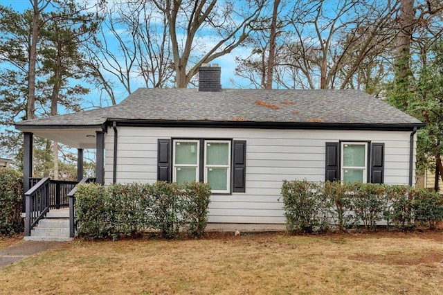 view of side of property with covered porch, roof with shingles, a yard, and a chimney