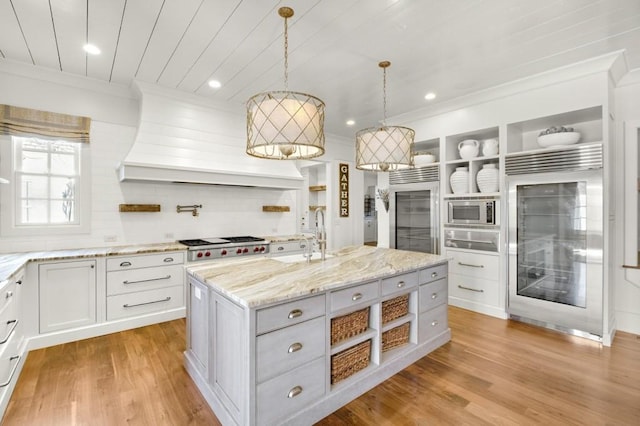 kitchen featuring built in appliances, premium range hood, a sink, open shelves, and a center island with sink