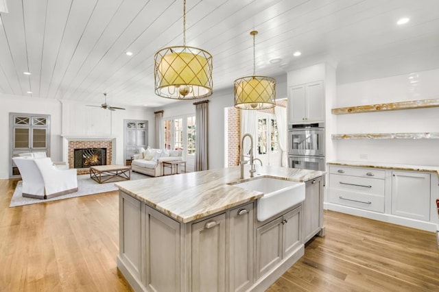 kitchen with stainless steel double oven, a sink, white cabinetry, hanging light fixtures, and an island with sink