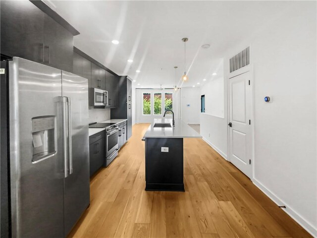 kitchen with appliances with stainless steel finishes, decorative light fixtures, light hardwood / wood-style flooring, sink, and an island with sink