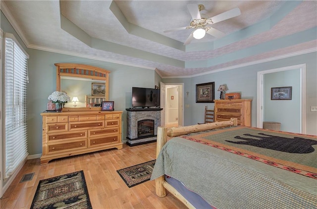 bedroom with a raised ceiling, a stone fireplace, crown molding, ceiling fan, and light hardwood / wood-style floors
