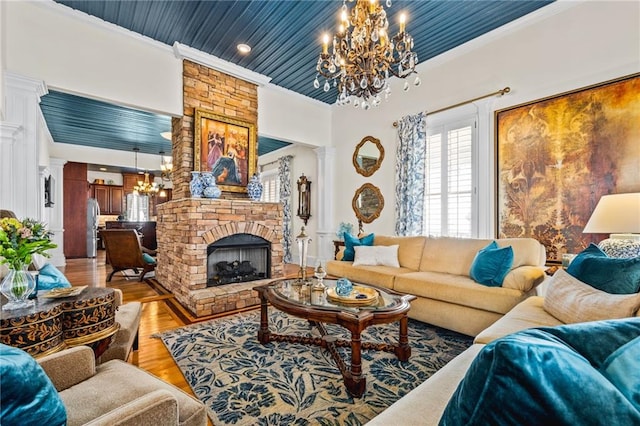 living room with ornate columns, light wood-style flooring, ornamental molding, a stone fireplace, and a chandelier