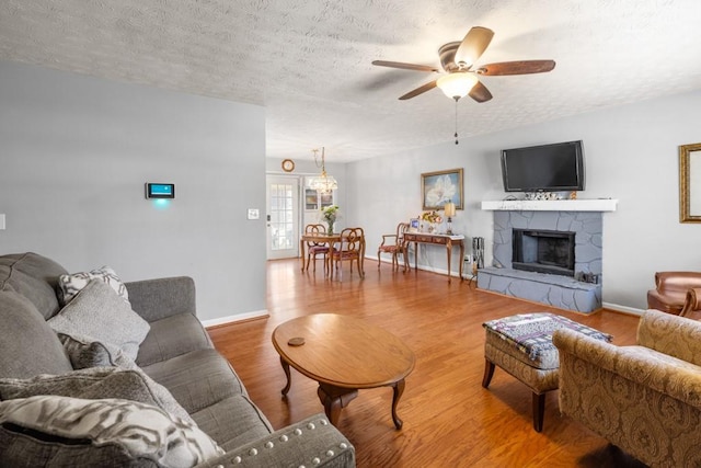 living room featuring baseboards, a stone fireplace, wood finished floors, a textured ceiling, and a ceiling fan