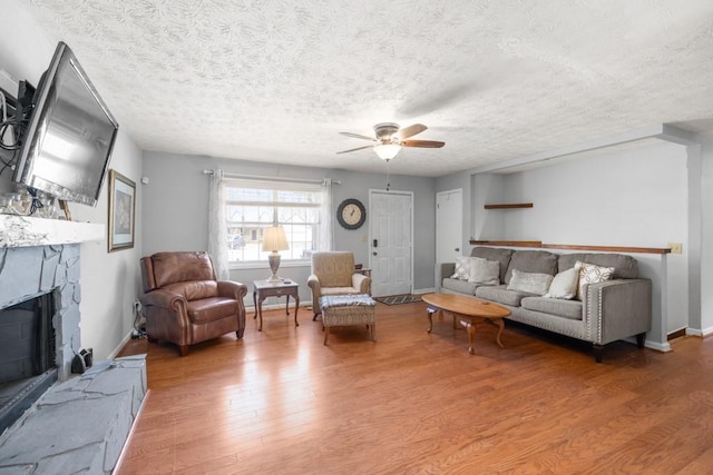 living area with light wood-type flooring, a ceiling fan, a textured ceiling, a stone fireplace, and baseboards