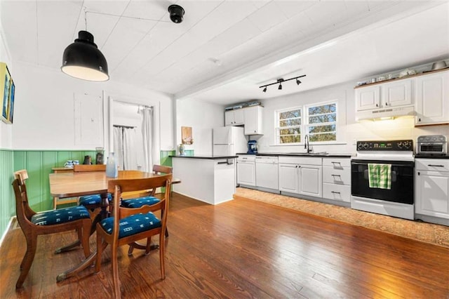 kitchen with dark countertops, under cabinet range hood, a wainscoted wall, wood finished floors, and white appliances