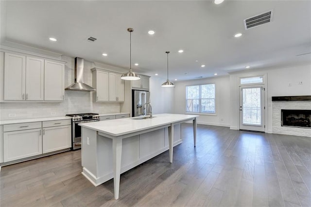 kitchen featuring wall chimney exhaust hood, stainless steel appliances, a kitchen island with sink, sink, and hanging light fixtures