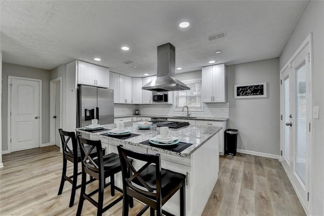 kitchen with appliances with stainless steel finishes, white cabinetry, island range hood, light stone countertops, and a kitchen bar