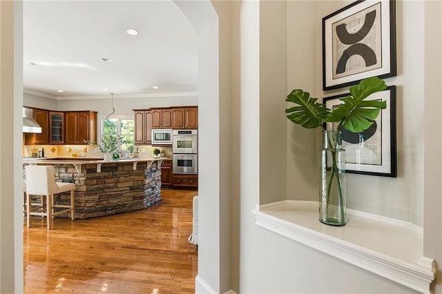 kitchen featuring appliances with stainless steel finishes, wood-type flooring, pendant lighting, wall chimney range hood, and ornamental molding