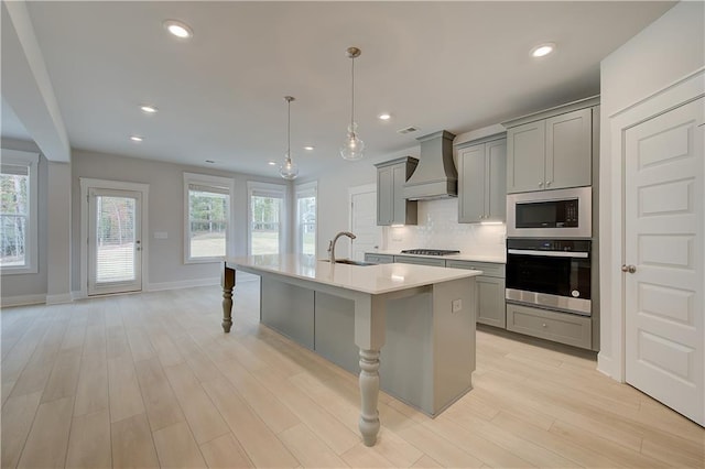 kitchen with gray cabinetry, custom range hood, stainless steel appliances, a kitchen island with sink, and light hardwood / wood-style floors