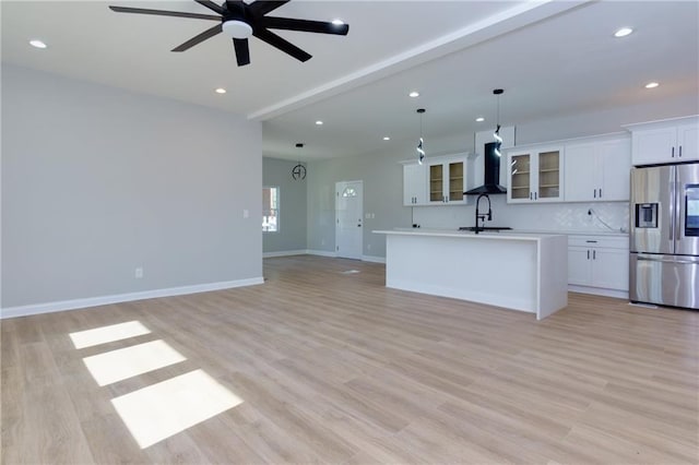 kitchen with wall chimney range hood, stainless steel fridge with ice dispenser, pendant lighting, a kitchen island with sink, and white cabinets