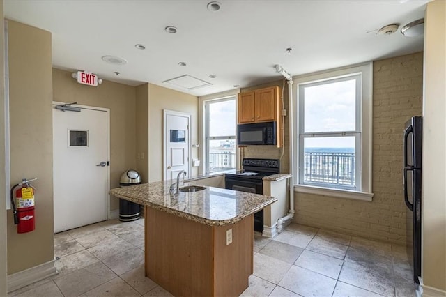 kitchen with sink, a center island with sink, brick wall, light stone countertops, and black appliances