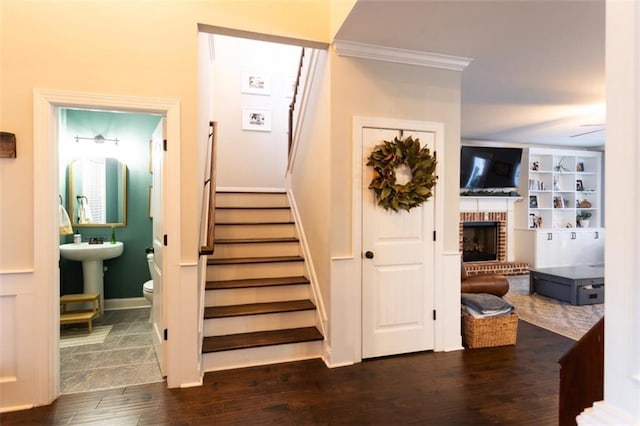 staircase featuring wood finished floors, a ceiling fan, baseboards, ornamental molding, and a brick fireplace