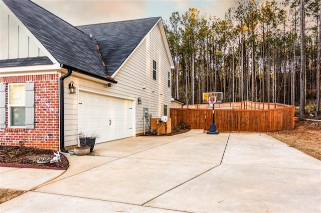 view of property exterior featuring concrete driveway, brick siding, roof with shingles, and fence