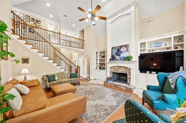 living room with a towering ceiling, ceiling fan, built in features, hardwood / wood-style floors, and a stone fireplace