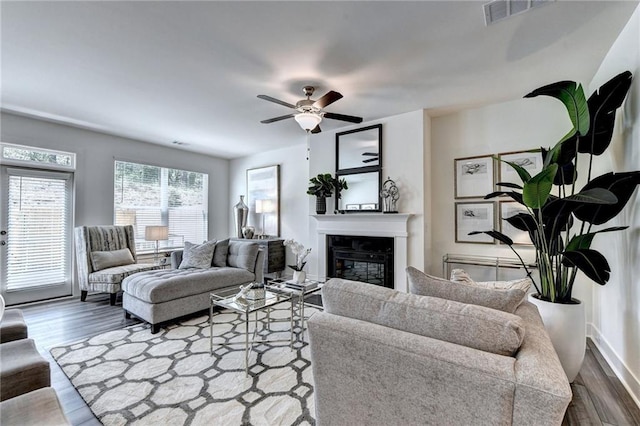 living area featuring a ceiling fan, a glass covered fireplace, visible vents, and dark wood finished floors