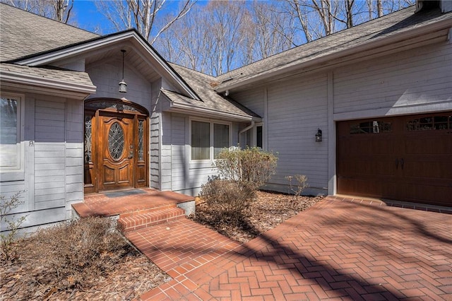 doorway to property featuring decorative driveway, a garage, and a shingled roof