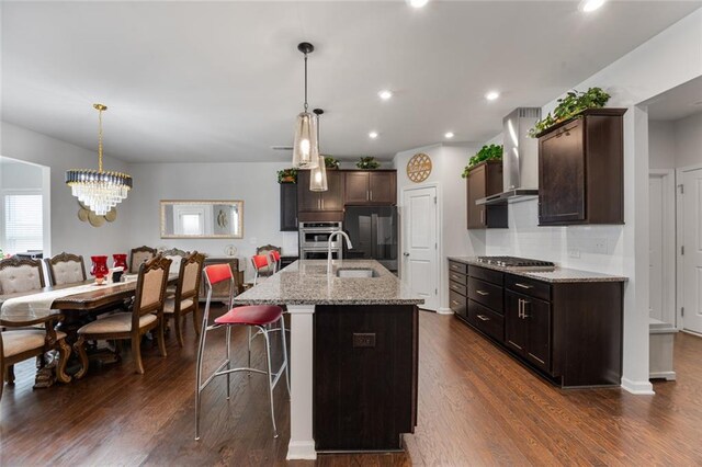 kitchen with pendant lighting, wall chimney range hood, stainless steel appliances, light stone counters, and an island with sink