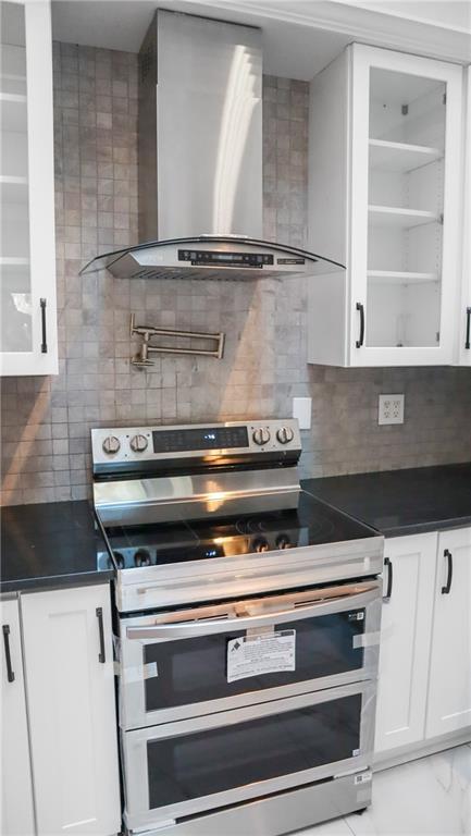 kitchen featuring wall chimney range hood, dark countertops, glass insert cabinets, and double oven range