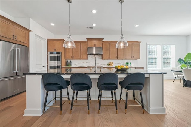 kitchen with under cabinet range hood, stainless steel appliances, visible vents, backsplash, and brown cabinets