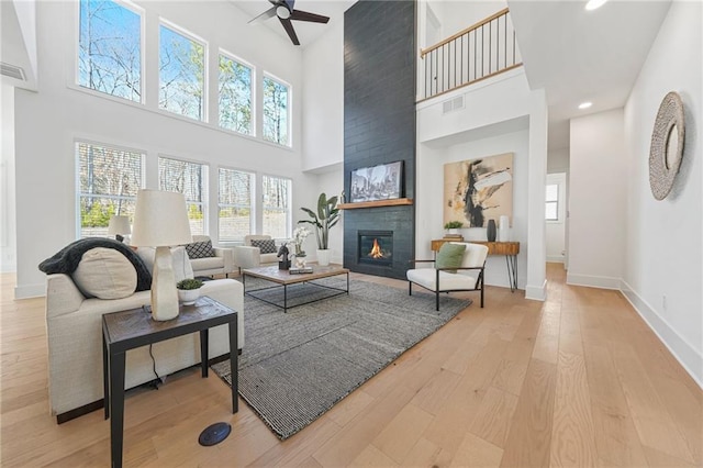 living room with ceiling fan, a tiled fireplace, light hardwood / wood-style flooring, and a towering ceiling