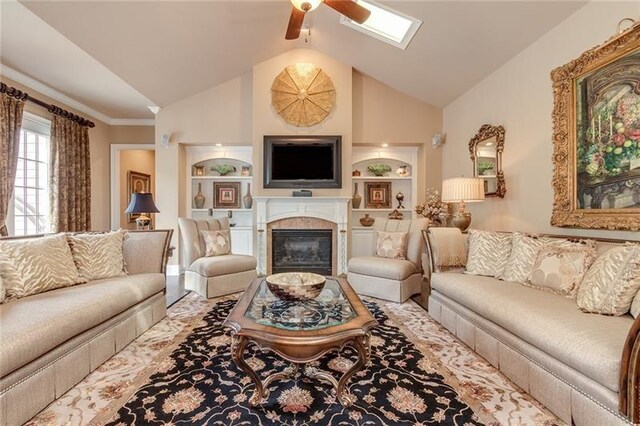 living area with built in shelves, vaulted ceiling with skylight, a glass covered fireplace, and a ceiling fan