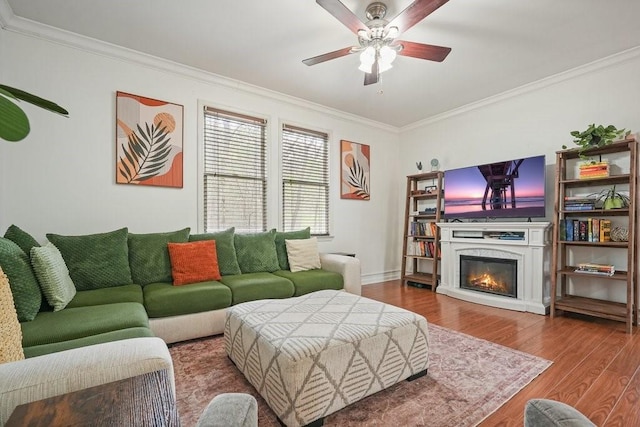 living room featuring a glass covered fireplace, a ceiling fan, baseboards, wood finished floors, and ornamental molding