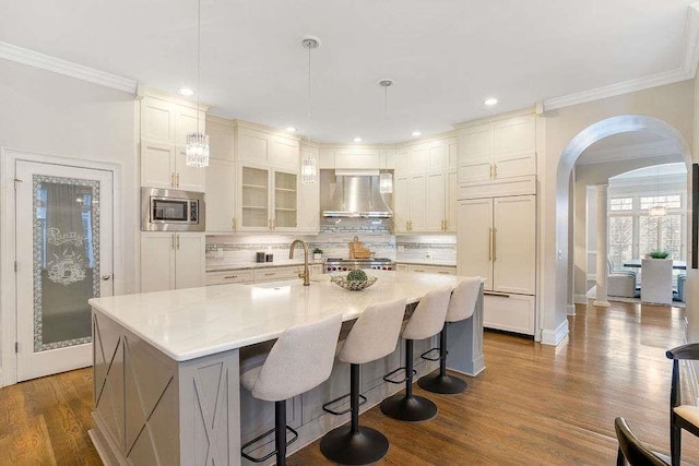 kitchen with built in appliances, wall chimney exhaust hood, a center island with sink, and sink