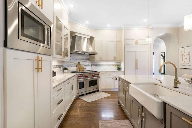 kitchen with sink, wall chimney exhaust hood, built in appliances, decorative light fixtures, and white cabinets