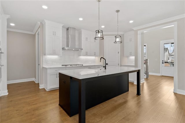 kitchen featuring wall chimney range hood, a center island with sink, light hardwood / wood-style floors, white cabinetry, and hanging light fixtures