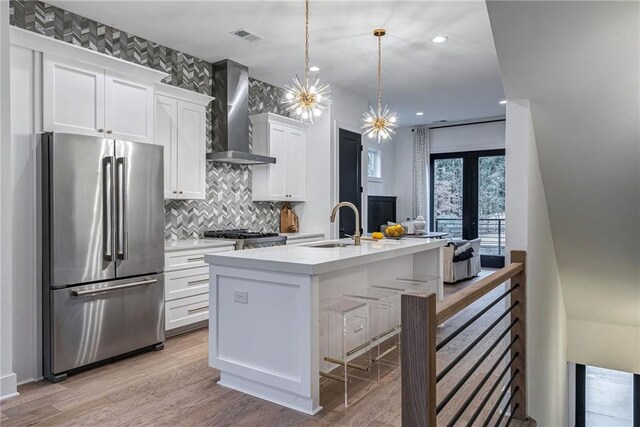 kitchen featuring stainless steel refrigerator, decorative light fixtures, white cabinets, wall chimney range hood, and a center island with sink