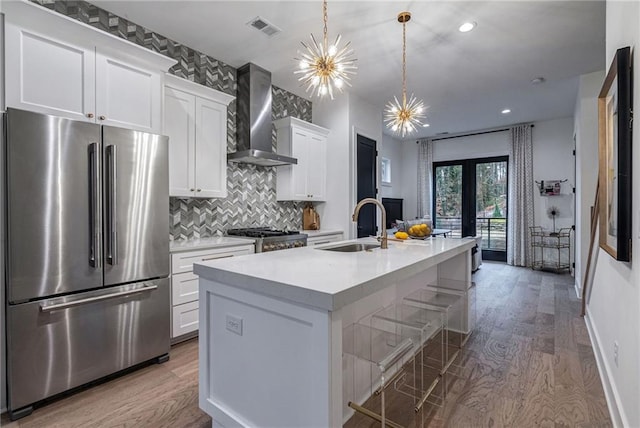 kitchen with wall chimney exhaust hood, white cabinetry, decorative light fixtures, stainless steel refrigerator, and an island with sink