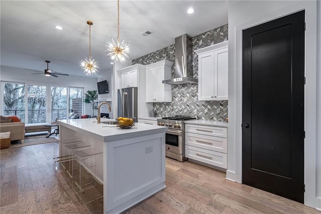 kitchen with white cabinets, premium appliances, hanging light fixtures, a center island with sink, and wall chimney exhaust hood