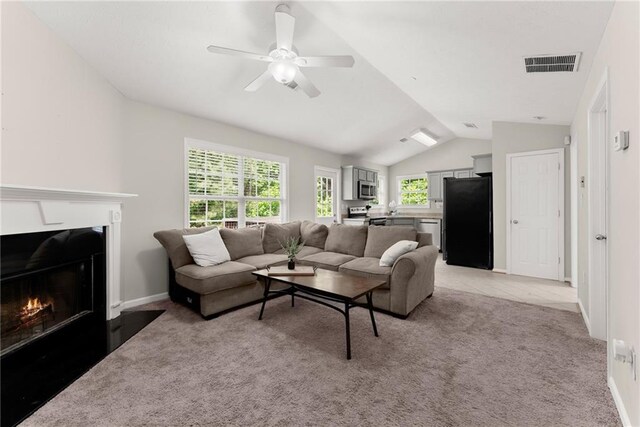 living room featuring a ceiling fan, light colored carpet, a fireplace with flush hearth, visible vents, and vaulted ceiling