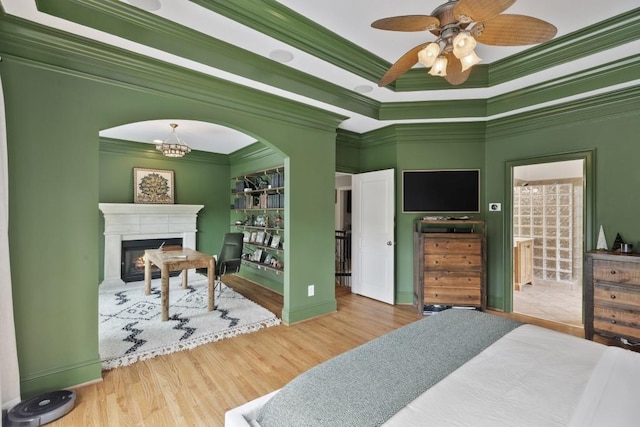 bedroom featuring ornamental molding, a glass covered fireplace, wood finished floors, baseboards, and a ceiling fan