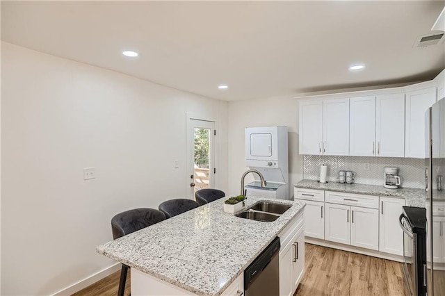 kitchen featuring stacked washing maching and dryer, stainless steel dishwasher, sink, an island with sink, and light wood-type flooring