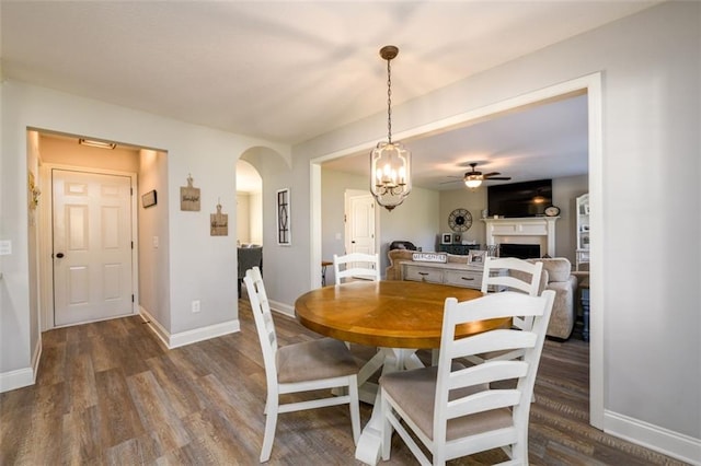 dining area with arched walkways, a fireplace, wood finished floors, baseboards, and ceiling fan with notable chandelier