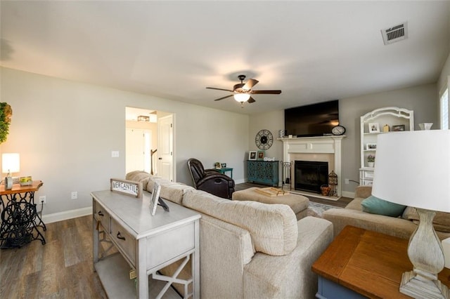 living area with a fireplace with raised hearth, dark wood-type flooring, visible vents, baseboards, and a ceiling fan