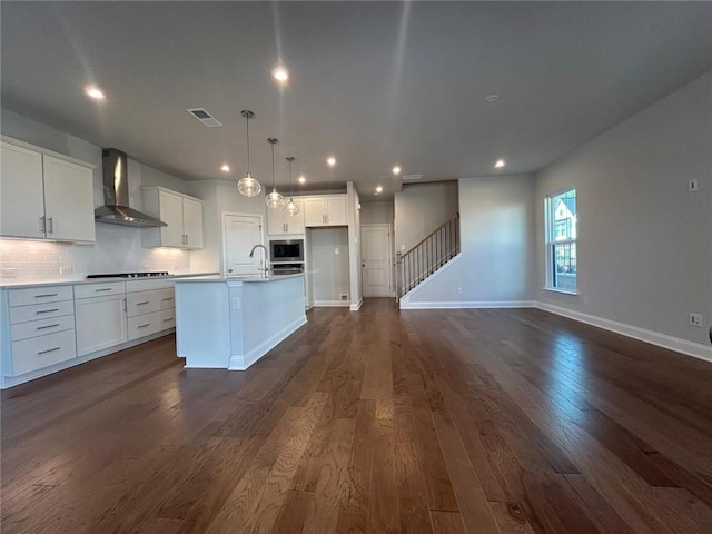 kitchen featuring a sink, wall chimney exhaust hood, stovetop, stainless steel microwave, and a center island with sink