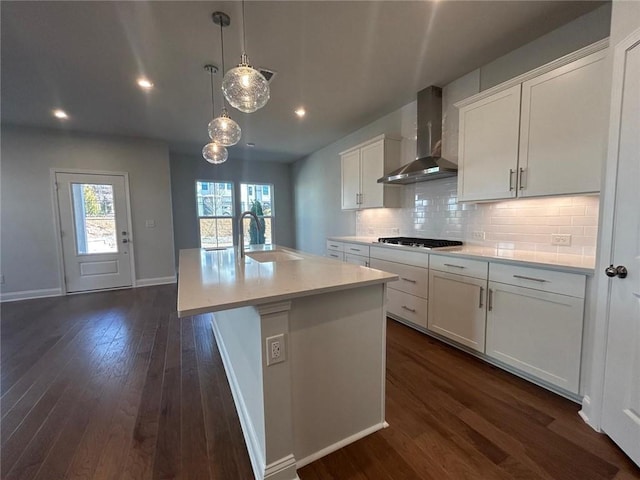 kitchen with dark wood-style floors, wall chimney exhaust hood, a sink, a kitchen island with sink, and backsplash