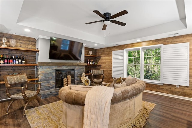 living room featuring a stone fireplace, a tray ceiling, dark hardwood / wood-style flooring, and wood walls