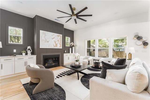 living area with plenty of natural light, light wood finished floors, visible vents, a glass covered fireplace, and ceiling fan