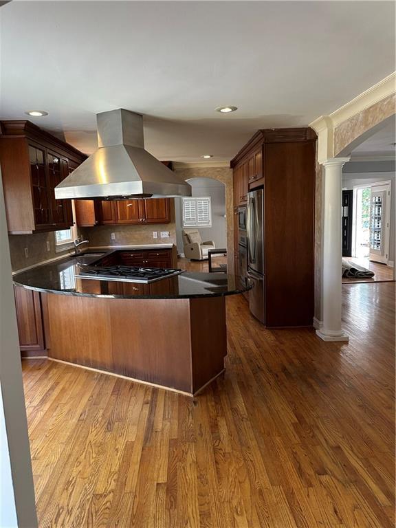 kitchen featuring island exhaust hood, wood finished floors, arched walkways, appliances with stainless steel finishes, and decorative columns