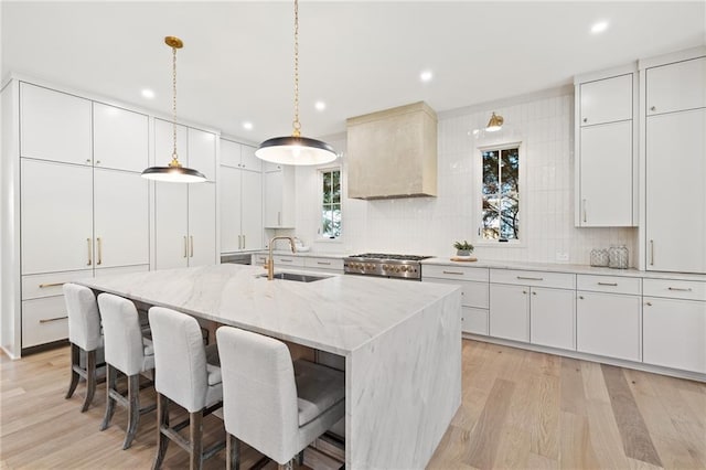 kitchen featuring sink, premium range hood, stove, an island with sink, and white cabinets