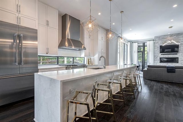 kitchen with wall chimney range hood, a fireplace, dark hardwood / wood-style flooring, white cabinetry, and stainless steel built in fridge