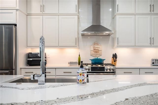 kitchen with light stone counters, stainless steel appliances, white cabinets, backsplash, and wall chimney exhaust hood