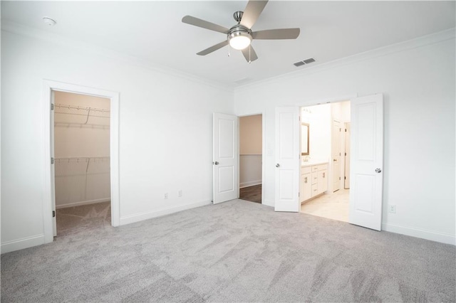 unfurnished bedroom with crown molding, ensuite bath, ceiling fan, a walk in closet, and light colored carpet