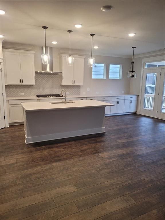 kitchen featuring white cabinets, wall chimney range hood, sink, hanging light fixtures, and an island with sink