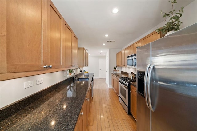 kitchen with light wood finished floors, stainless steel appliances, dark stone counters, and a sink